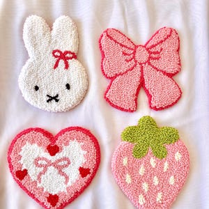 Può includere: Quattro sottobicchieri fatti a mano e strutturati in varie forme. Include un coniglietto bianco con un fiocco rosso, un fiocco rosa, un cuore rosa con un fiocco e cuori rossi e una fragola rosa con una parte superiore verde e semi bianchi.