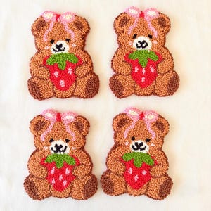 May include: Four brown teddy bear-shaped decorative items, each holding a red strawberry with green leaves. The bears have pink bows and black facial features. The items are made of a textured material, possibly yarn or fabric.
