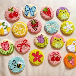 May include: A collection of colorful, round, handmade coasters with various designs. The coasters feature images of fruits like cherries, strawberries, and lemons, along with a bow, a flower, a bee, and a frog. The coasters are made with a textured, looped yarn.