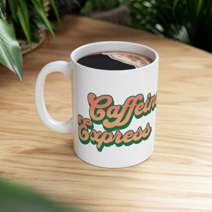 Puede incluir: Taza de cerámica blanca con un diseño verde, naranja y amarillo que dice "Caffeine Express". La taza está llena de café negro.