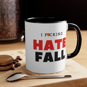 May include: White coffee mug with a black rim and handle. The mug has black text that says "I F*CKING HATE FALL" with a red maple leaf in the word "F*CKING".