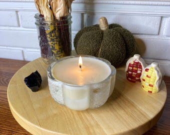 May include: A lit candle in a clear, pumpkin-shaped glass container sits on a round wooden tray. Nearby are a glass jar filled with dried corn, a textured olive green fabric pumpkin, two small corn-shaped ceramic shakers, and a black stone.