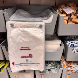 May include: A white plastic bag with the text "little bag, lots of extras" and the Chick-fil-A logo in red. The bag is hanging from a metal hook on a gray plastic bin. The bin is filled with white paper napkins. There are other gray bins filled with various items, including small packets of sauces and condiments.