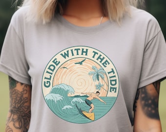 Puede incluir: Una camiseta gris con un gráfico de estilo vintage de un surfista montando una ola. El gráfico dice "Glide with the Tide" en un círculo con palmeras y una puesta de sol.