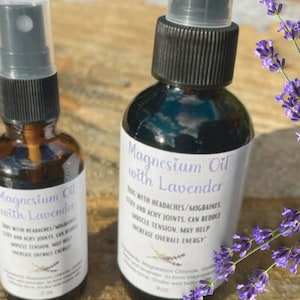 May include: Two amber glass bottles with black spray tops and white labels. The labels read "Magnesium Oil with Lavender" and list benefits including relief from headaches, migraines, sore and achy joints, and muscle tension. The bottles are sitting on a wooden surface with a sprig of lavender flowers in the background.