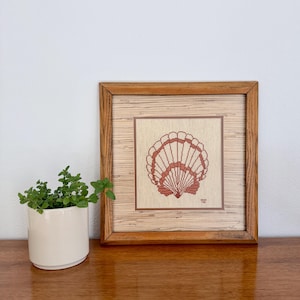 May include: A framed cross-stitch of a seashell in shades of red and tan. The artwork is displayed in a wooden frame with a light-colored, textured background. A small potted plant sits to the left of the frame.