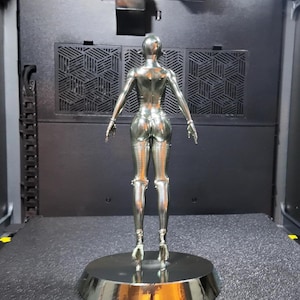 May include: A chrome-colored sculpture of a female figure, viewed from the back. The figure is posed with arms outstretched, standing on a circular base. The sculpture has a smooth, reflective surface and is set against a dark background.