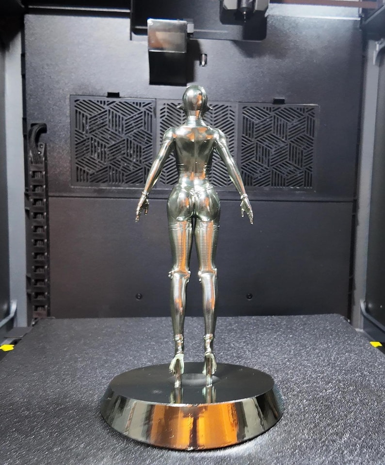 May include: A chrome-colored sculpture of a female figure, viewed from the back. The figure is posed with arms outstretched, standing on a circular base. The sculpture has a smooth, reflective surface and is set against a dark background.
