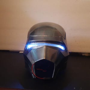Puede incluir: Un casco plateado con una visera negra y luces azules. El casco tiene un diseño futurista y está hecho de metal.