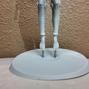 May include: A light gray doll stand with two legs and feet attached to a circular base. The legs are connected to the base by metal rods. The background is a textured brown wall.