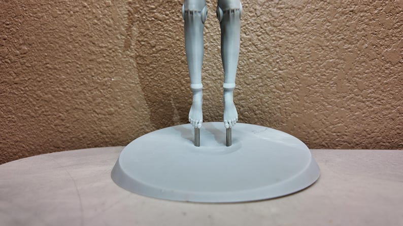 May include: A light gray doll stand with two legs and feet attached to a circular base. The legs are connected to the base by metal rods. The background is a textured brown wall.