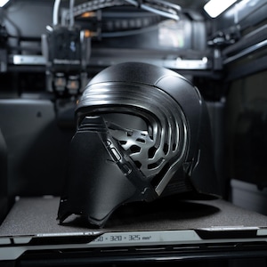 以下が含まれることがあります： スターウォーズのキャラクター、カイロ・レンの黒いヘルメットが3Dプリンター内に展示されています。ヘルメットは、メタリックシルバーのフェイスマスクとマットブラック仕上げの独特のデザインです。プリンターは暗い環境にあります。