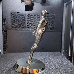 May include: A chrome-colored, stylized sculpture of a female figure, posed in a dynamic stance. The figure is mounted on a circular base. The sculpture has articulated joints and a smooth, reflective surface, suggesting a futuristic or robotic theme.
