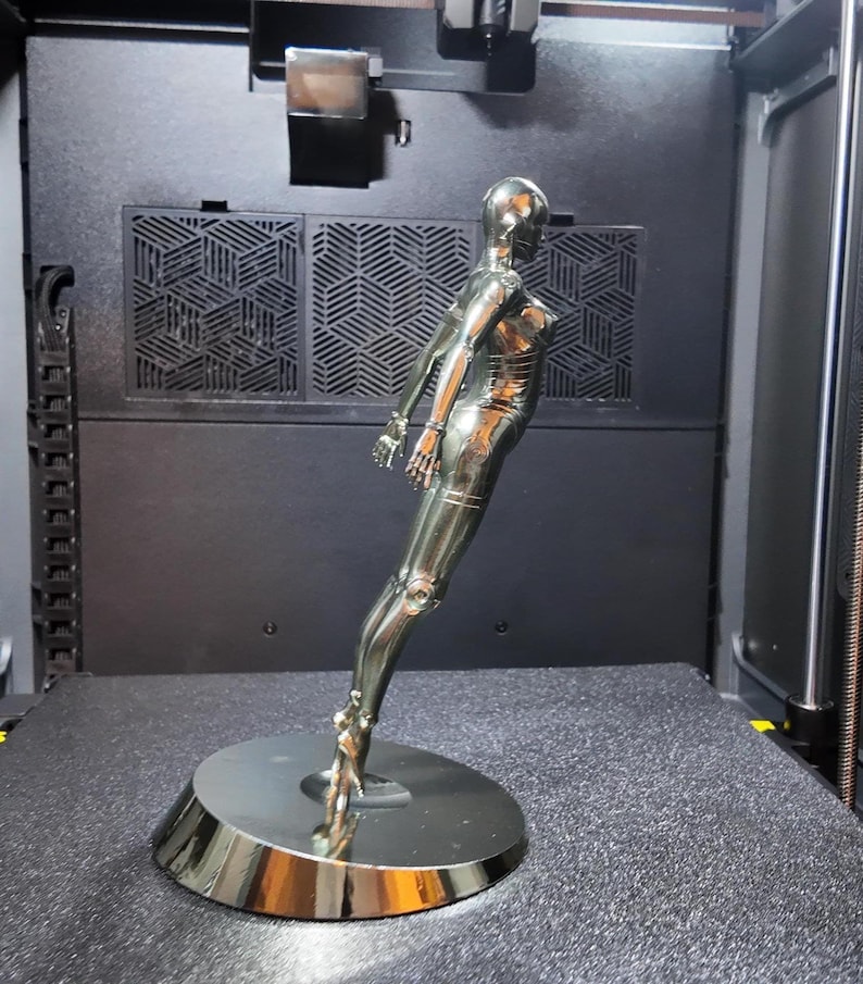 May include: A chrome-colored, stylized sculpture of a female figure, posed in a dynamic stance. The figure is mounted on a circular base. The sculpture has articulated joints and a smooth, reflective surface, suggesting a futuristic or robotic theme.