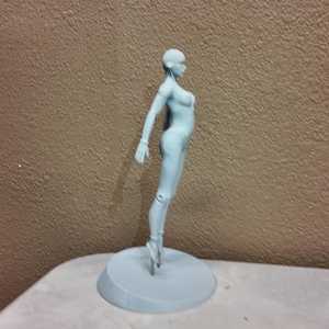 May include: A light blue figurine of a person in a ballet pose. The figure is standing on a round base. The background is a textured brown wall.