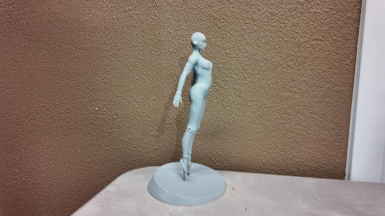 May include: A light blue figurine of a person in a ballet pose. The figure is standing on a round base. The background is a textured brown wall.