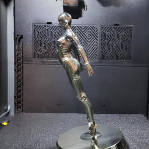 May include: A chrome-colored sculpture of a female figure, posed in a dynamic stance. The figure is mounted on a circular base. The sculpture has a sleek, metallic finish, and is set against a dark background.