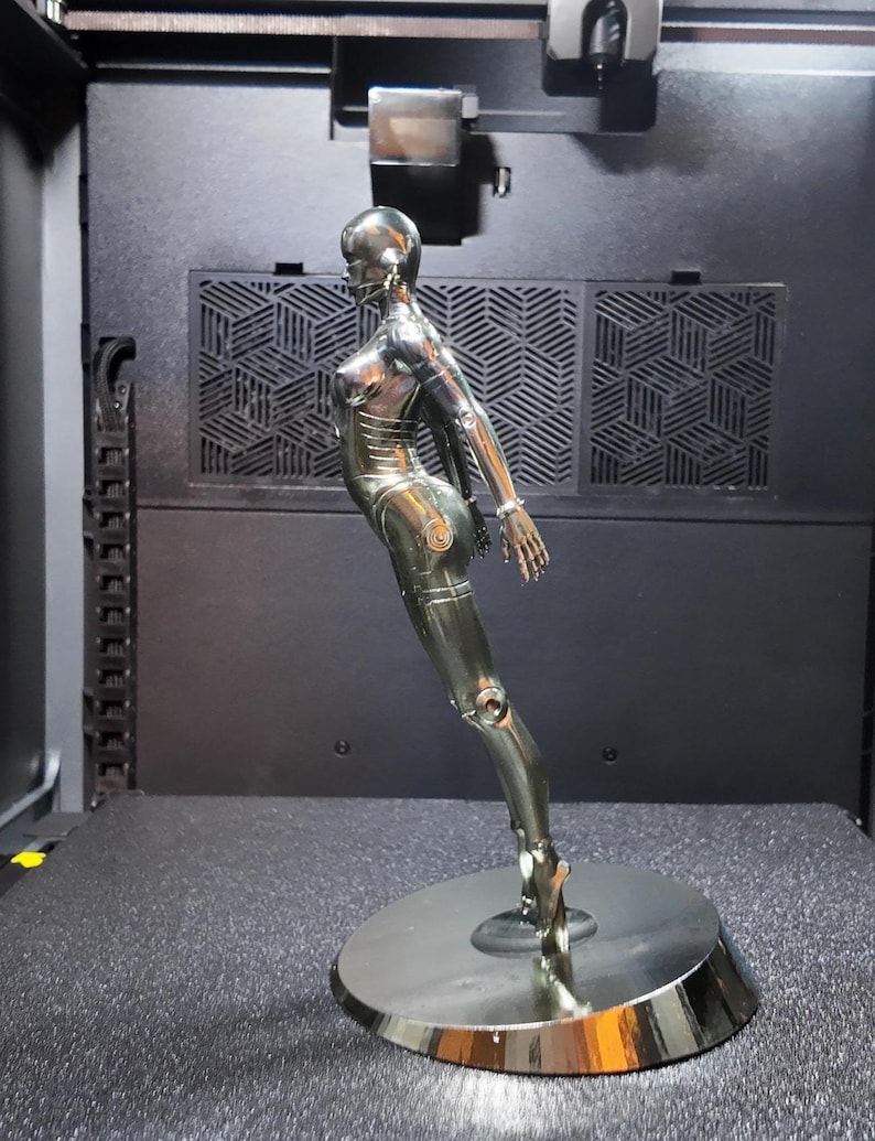 May include: A chrome-colored sculpture of a female figure, posed in a dynamic stance. The figure is mounted on a circular base. The sculpture has a sleek, metallic finish, and is set against a dark background.