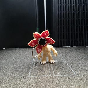Puede incluir: Un llavero Demogorgon con una cabeza roja en forma de flor con dientes blancos y un cuerpo beige. El llavero es de plástico y tiene un anillo plateado. La figura está sobre una superficie gris.