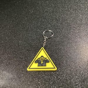 Könnte beinhalten: Ein gelber, dreieckiger Schlüsselanhänger mit schwarzem Rand und einer schwarzen Grafik einer Person, die eine Jacke und eine Halskette trägt. Der Schlüsselanhänger ist an einer silbernen Kette und einem Ring befestigt.