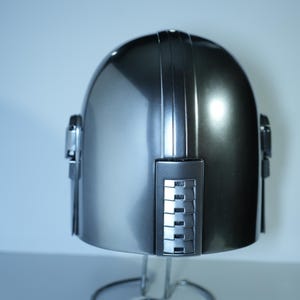May include: A metallic silver helmet with a rounded top and vertical ridges. The helmet has rectangular side pieces and is displayed on a silver stand. The helmet's surface reflects light, creating highlights and shadows.