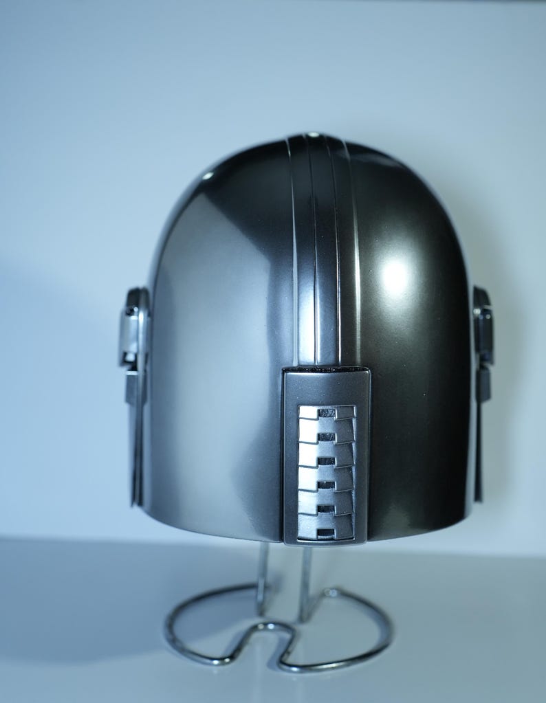 May include: A metallic silver helmet with a rounded top and vertical ridges. The helmet has rectangular side pieces and is displayed on a silver stand. The helmet's surface reflects light, creating highlights and shadows.