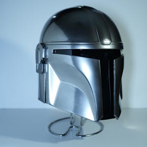 May include: A metallic silver helmet with a black visor, displayed on a wire stand. The helmet has a smooth, reflective surface with angular features and a distinct design. The helmet is a replica of a character's helmet from a popular science fiction franchise.