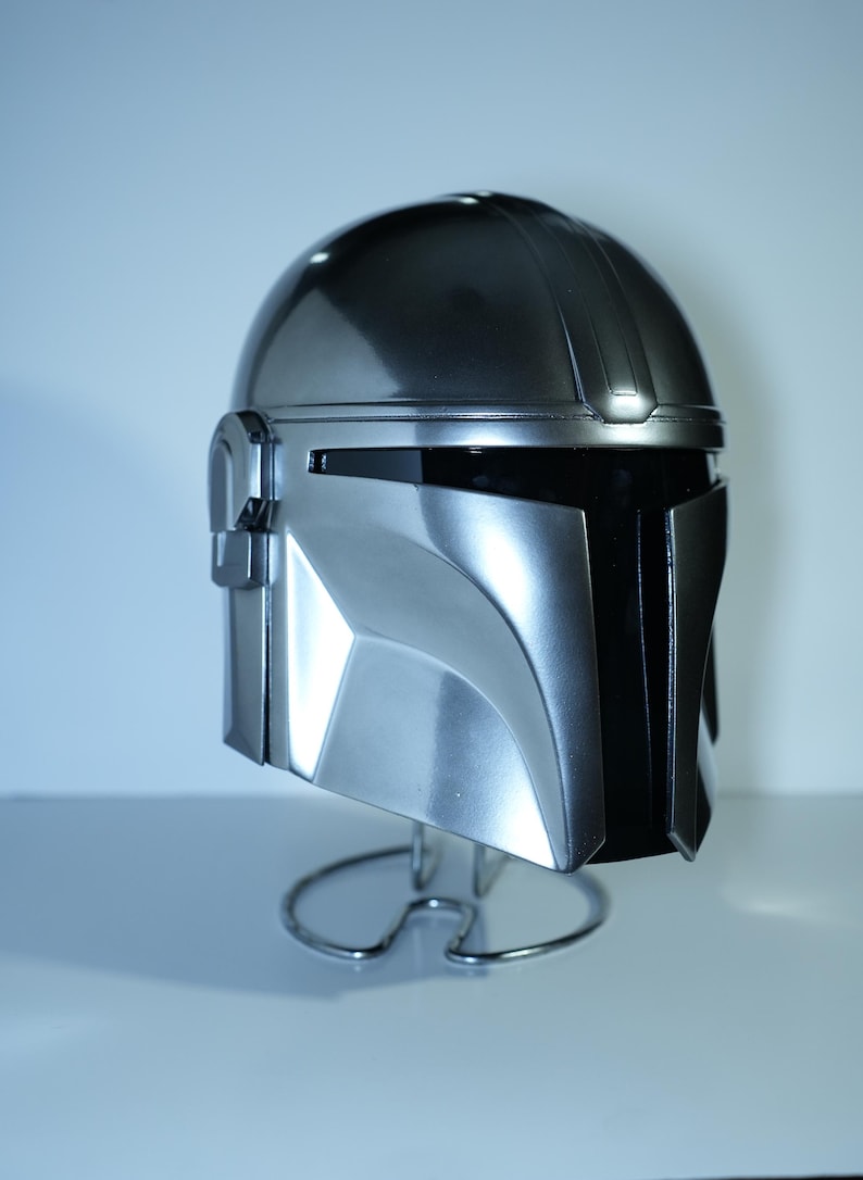 May include: A metallic silver helmet with a black visor, displayed on a wire stand. The helmet has a smooth, reflective surface with angular features and a distinct design. The helmet is a replica of a character's helmet from a popular science fiction franchise.