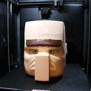 Op de afbeelding: Een 3D-geprint model van het hoofd van een cartoonfiguur. Het hoofd is beige met bruine wenkbrauwen en gele ogen. Het hoofd staat op een zwart oppervlak in een 3D-printer.