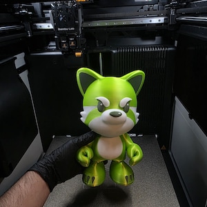 May include: A 3D-printed figurine of a cartoon fox with a green and white color scheme. The fox has a stern expression and is held in front of a 3D printer. The figurine is approximately 6 inches tall.