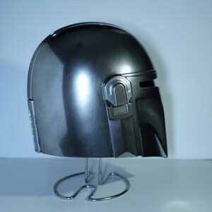 May include: A metallic gray helmet with a sleek, curved design. The helmet features a visor and detailed side panels. It is displayed on a silver metal stand, showcasing its polished surface and overall shape.