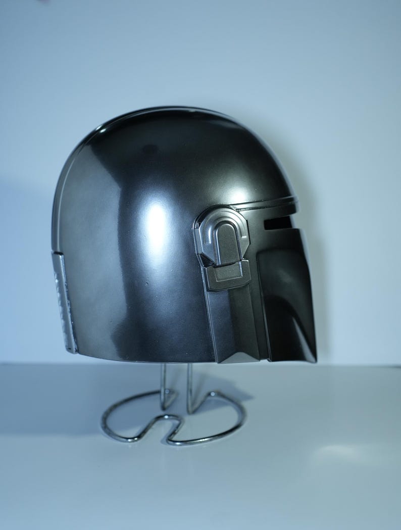 May include: A metallic gray helmet with a sleek, curved design. The helmet features a visor and detailed side panels. It is displayed on a silver metal stand, showcasing its polished surface and overall shape.