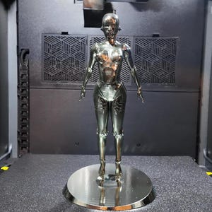 Puede incluir: Una figura de robot plateado metálico, de pie sobre una base circular. El robot tiene una forma humanoide con detalles, incluyendo cabeza, torso, brazos y piernas. La figura está sobre un fondo oscuro.