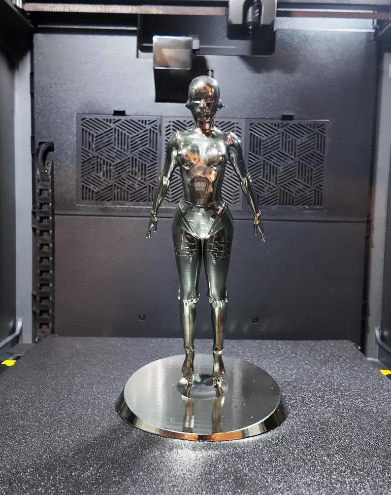 May include: A metallic silver robot figurine, standing on a circular base. The robot has a humanoid form with detailed features, including a head, torso, arms, and legs. The figurine is set against a dark background.