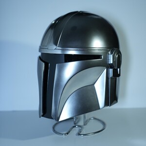 May include: A metallic silver Mandalorian helmet with a black visor and distinctive angular design. The helmet is displayed on a silver wire stand, showcasing its sleek, futuristic aesthetic. The helmet's surface reflects light, highlighting its detailed construction.