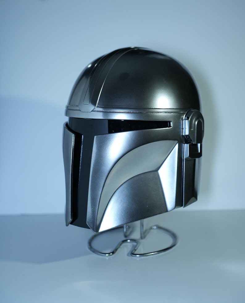 May include: A metallic silver Mandalorian helmet with a black visor and distinctive angular design. The helmet is displayed on a silver wire stand, showcasing its sleek, futuristic aesthetic. The helmet's surface reflects light, highlighting its detailed construction.