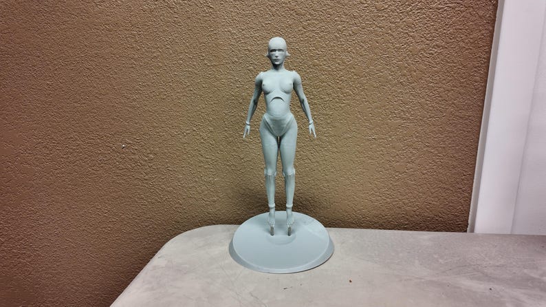 May include: A light gray, stylized human figure, standing on a circular base. The figure has a smooth, minimalist design, with articulated joints and a neutral expression. The figure is approximately 12 inches tall.