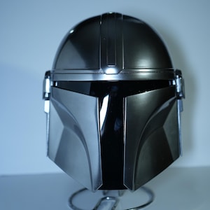 May include: A metallic silver Mandalorian helmet, featuring a sleek, angular design. The helmet has a dark visor and is displayed on a silver wire stand. The helmet's surface reflects light, highlighting its smooth, polished finish.