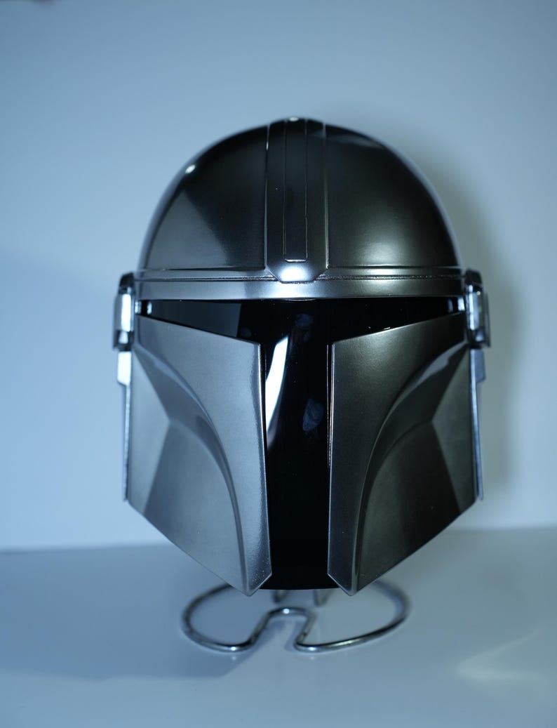 May include: A metallic silver Mandalorian helmet, featuring a sleek, angular design. The helmet has a dark visor and is displayed on a silver wire stand. The helmet's surface reflects light, highlighting its smooth, polished finish.