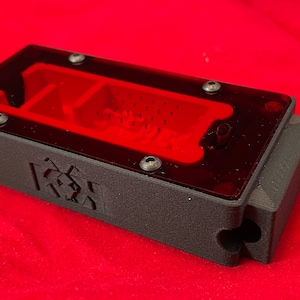 May include: A black 3D printed ant farm with a clear red plastic top. The farm has a small opening on the side and a logo on the bottom that says "[K3X]"