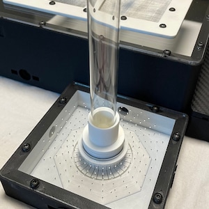 May include: A black square box with a white interior and a clear glass tube. The tube is inserted into a white plastic base with a circular pattern of small holes. The box is surrounded by a white surface.