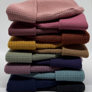 May include: A stack of folded beanie hats in various colors including dusty pink, magenta, rust, mustard, tan, sage green, purple, teal, navy, and charcoal. The text Organic Cotton Beanie Hats and WINDBORNE CREATIONS T-SHIRTS & MORE is visible.