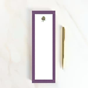 May include: A rectangular notepad with a purple and white striped border and a white center panel featuring a small green artichoke illustration at the top, accompanied by a slim gold-colored metal pen placed vertically to the right side.