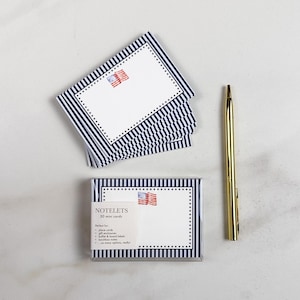 May include: A set of 30 mini notecards with a patriotic theme. The cards feature a white background with a small American flag and a navy blue and white striped border. A gold pen rests beside the cards.