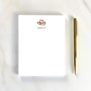 May include: A white notepad with a watercolor illustration of a cowboy hat and the words "Giddy Up" in brown. A gold pen rests beside the notepad. The notepad and pen are on a white surface.