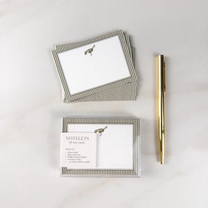 May include: A stack of white notelets with a thin brown striped border and a small bird illustration, accompanied by a packaged set of 30 mini cards featuring the same design and a gold-toned metal pen. The packaging includes the text NOTELETS 30 mini cards.