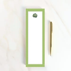 May include: A rectangular notepad with a white center, a green and white striped border, and a small green cabbage illustration at the top, accompanied by a slim gold-colored metal pen positioned vertically to the right of the notepad.