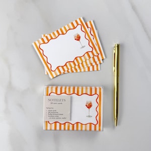 May include: A set of "NOTELETS" mini cards with an orange and white striped design and a watercolor illustration of a cocktail glass. A gold pen rests beside the cards. The cards are packaged in a clear plastic box.