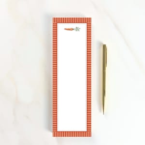 May include: A rectangular notepad with a white center and a border featuring orange and white vertical stripes. A small illustration of a single orange carrot with green leafy tops is positioned at the top center. A gold metal pen lies beside it.