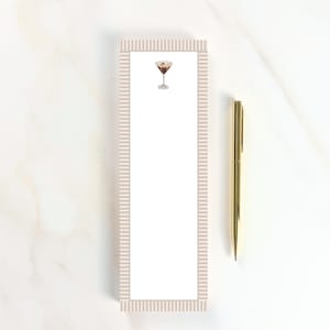 May include: A rectangular notepad with a light beige striped border and a cocktail illustration at the top. A gold pen rests beside the notepad. The notepad is white with a cocktail illustration.